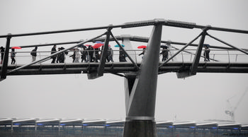 Millenium bridge umbrellas This urban architecture photograph captures the Millennium Bridge in London, United Kingdom, during a rainy afternoon in winter. The main subject of the image is the Millennium Bridge, which is seen carrying numerous pedestrians, many of whom are holding umbrellas as they cross. The scene highlights the distinctive design of the bridge and its role as a key architectural landmark in central London, with the overcast sky and muted light typical of winter afternoons. The backdrop includes modern city infrastructure, further emphasizing the urban setting of this London bridge.